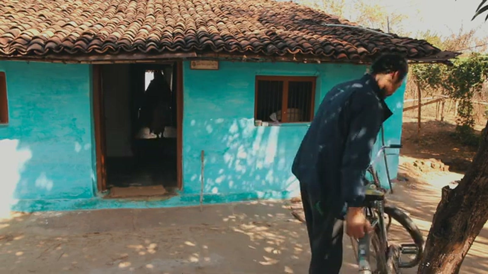 A man with a bicycle outside a turquoise village house, a frame from Aas