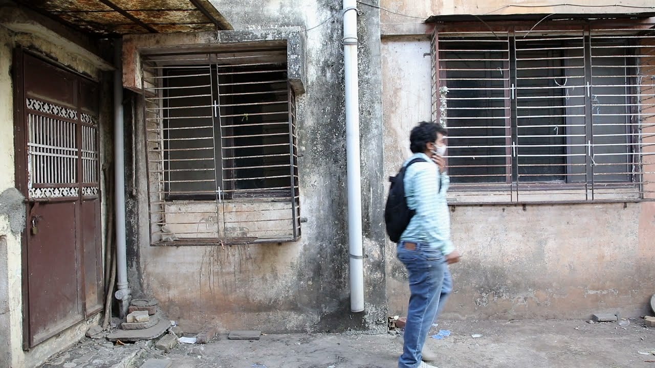 A man in a mask and backpack walks past a weathered building with barred windows