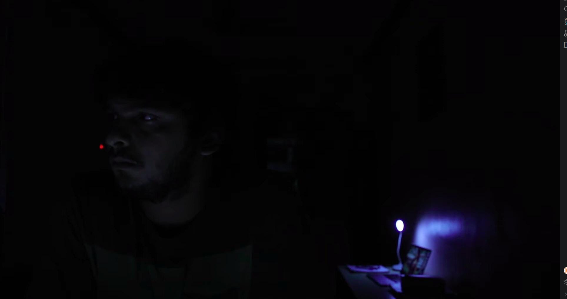 A man sits in a nearly dark room, his face caught by a small lamp