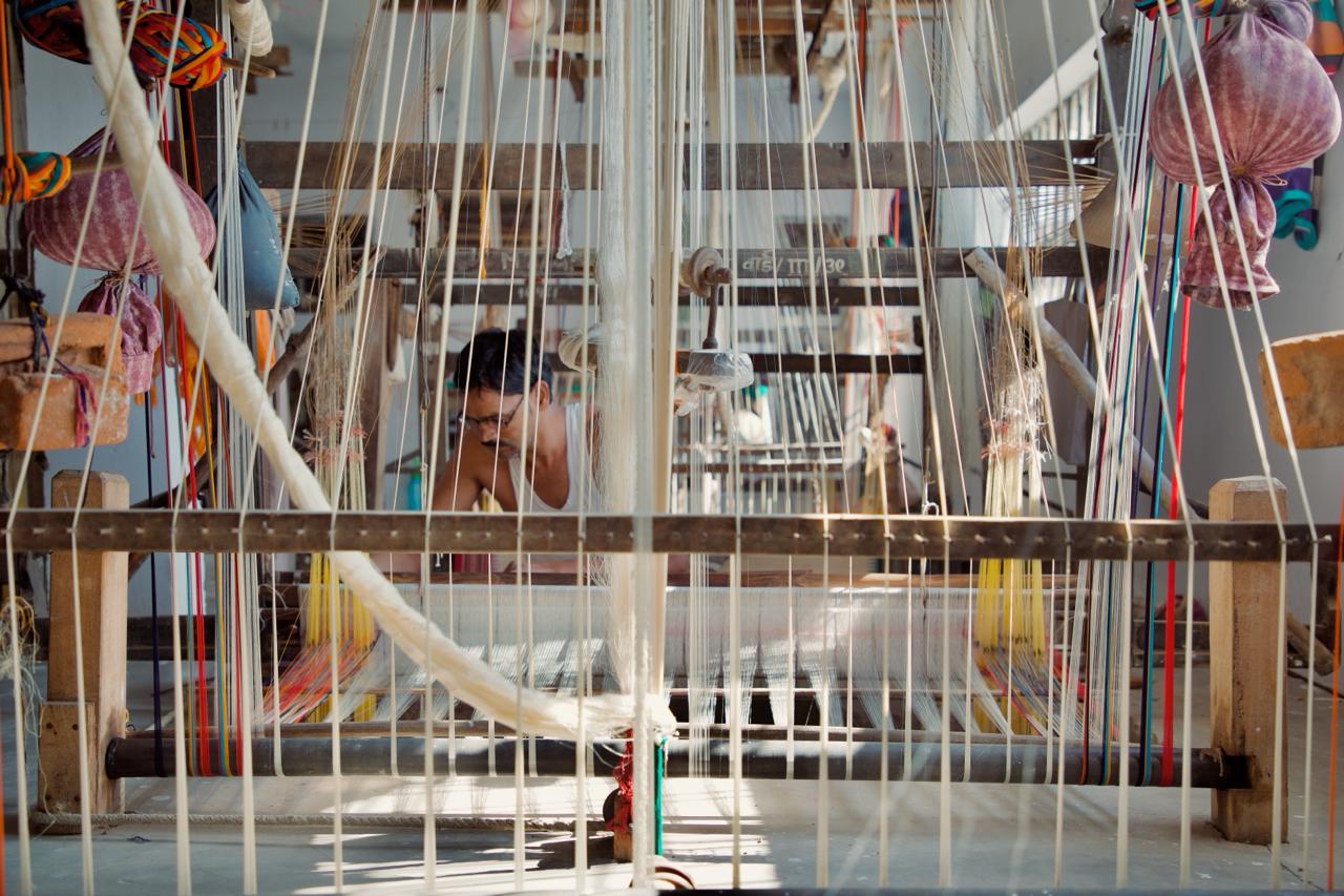 A weaver at a wooden handloom, seen through hanging warp threads
