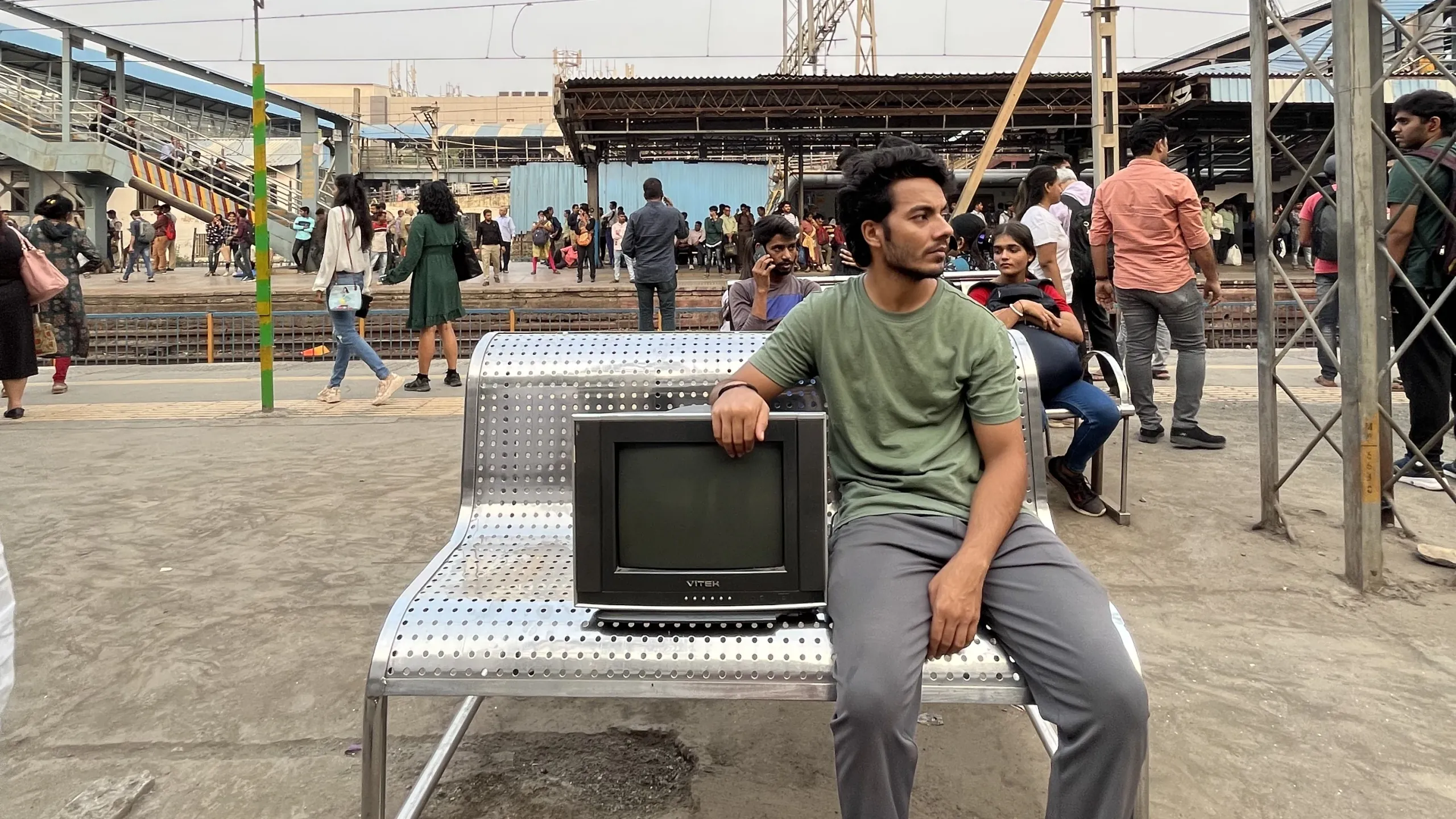 A young actor sits beside the television at a crowded railway station
